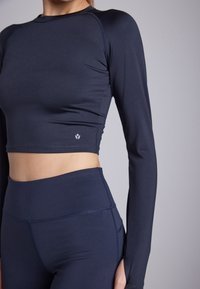 Tamno plavi dugi rukavi crop top s glatkom teksturom i prilagođenim dizajnom, u kombinaciji s tajicama visokog struka, koje imaju suptilne detalje šivanja.