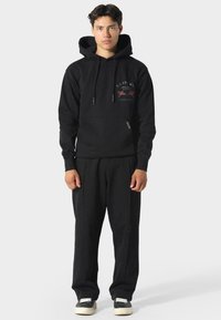 Sudadera negra con un bolsillo tipo canguro en la parte frontal, capucha con cordones y detalles de logo. Combinada con pantalones de chándal negros de corte holgado y zapatillas.
