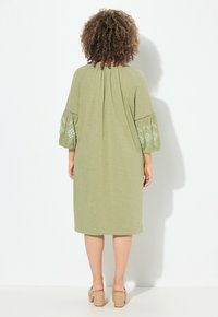 Oliwkowa sukienka z plisowanym tyłem i haftowanymi, balonowymi rękawami. Średniej długości, z miękkiego materiału, noszona z lekkimi, grubymi sandałami na obcasie.