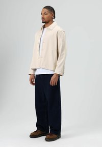 Beige jack met knopen over een wit t-shirt, gecombineerd met donkerblauwe, loszittende spijkerbroek en bruine wandelschoenen. Effen achtergrond.