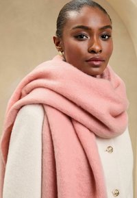 Pehme roosakas villane sall on kaela ümber käinud, kombineeritud kreemivärvi mantliga, millel on kuldsed nööbid. Peen tekstuur on nähtav.