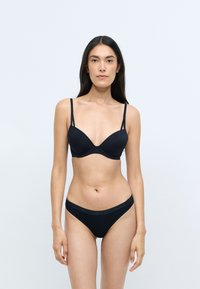 Soutien-gorge noir et culotte assortie en tissu lisse. Le soutien-gorge présente un décolleté plongeant et des fines bretelles ; la culotte possède une bande logo à la taille.