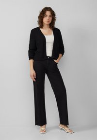 Schwarze gestrickte Strickjacke mit weitem Ausschnitt, über einem weißen Tanktop getragen, kombiniert mit schwarzen hochgeschnittenen Jeans und weißen Sandalen mit Riemchen.