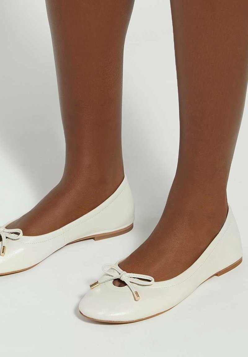 Jambes à la peau foncée portant des ballerines en cuir blanc avec de petits nœuds et des détails de lacets dorés, debout sur un sol blanc.