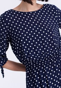 Námořnický modrý šaty mají bílé puntíky, krátké rukávy a elastický pas, vyrobené z měkké tkaniny pro pohodlí.