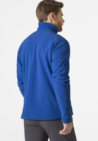 Niebieska polarowa kurtka z wysokim kołnierzem, z długimi rękawami i gładką teksturą. Krój obejmuje prosty dół oraz minimalne detale.