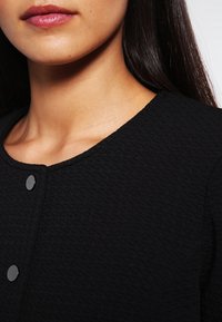 Tissu noir texturé avec un col rond et deux boutons-pression argentés. Vue rapprochée mettant en valeur le motif du tissu et sa finition lisse.