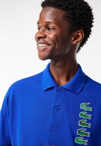 Polo bleu à manches courtes, avec une patte à trois boutons et cinq logos crocodile verts brodés sur le côté gauche de la poitrine.