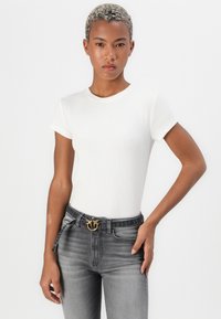 T-shirt bianca a costine con maniche corte, abbinata a jeans grigi scuri. I jeans presentano una fibbia decorativa dorata e un leggero effetto sbiadito.