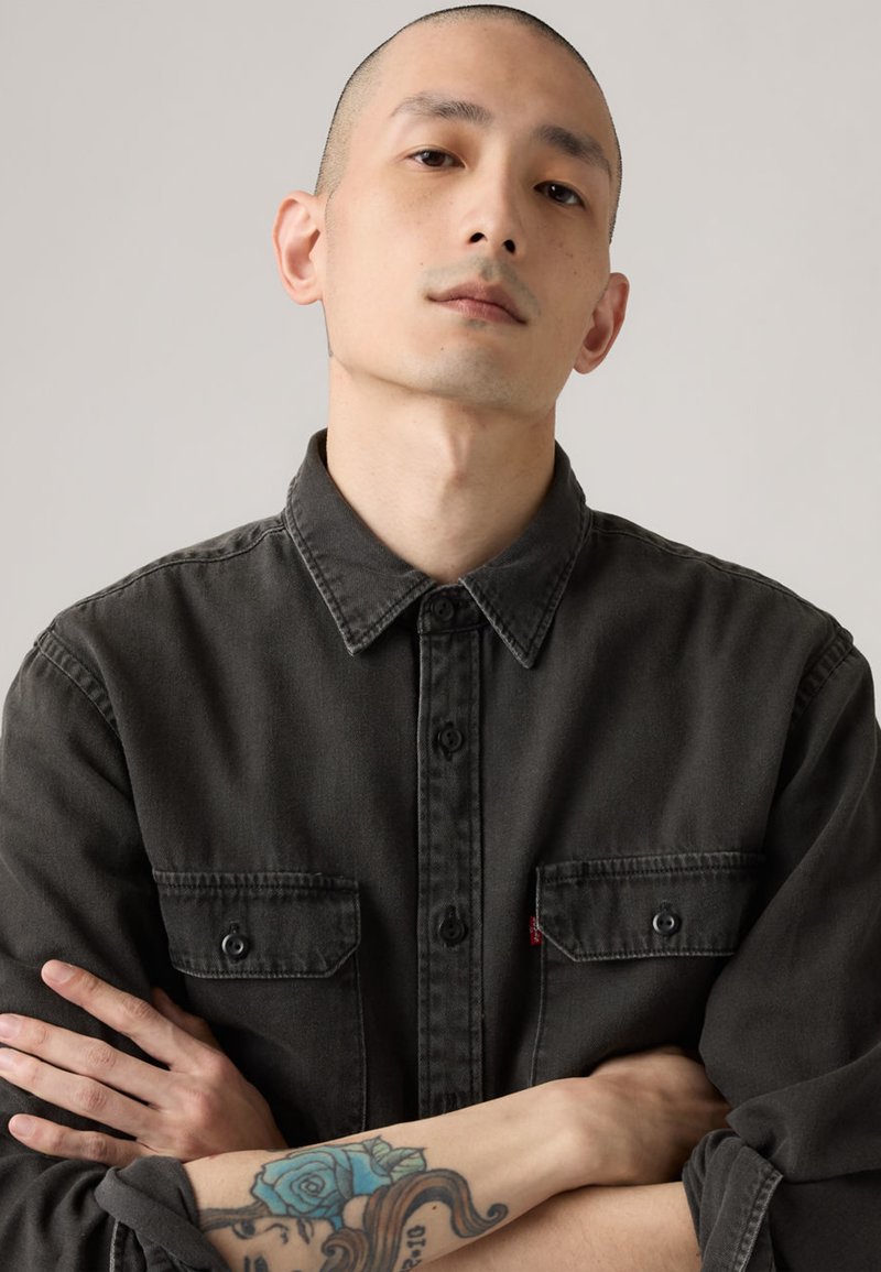 Chemise boutonnée gris foncé avec col, deux poches avant et une étiquette Levi's. Texture lisse, bras croisés, avec un tatouage sur l'avant-bras.