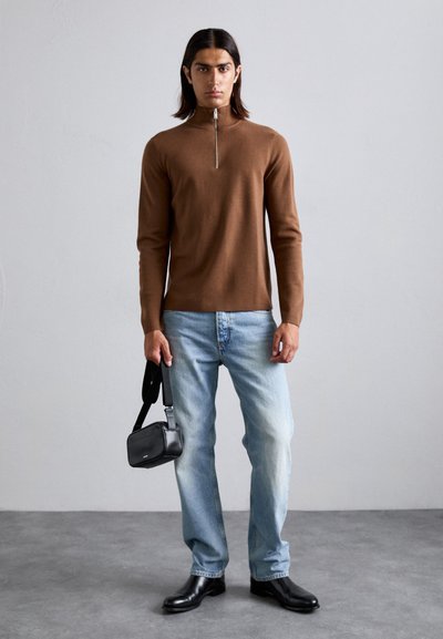 Mann in braunem Reißverschluss-Pullover, hellblauen Jeans, schwarzen Schuhen und mit einer kleinen schwarzen Schultertasche, steht auf einem grauen Boden vor einer weißen Wand.