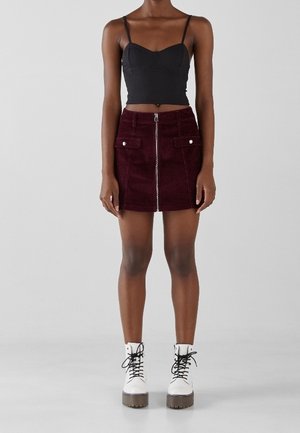 Kvinde iført sort cropped tanktop, bordeaux cordnede mininederdel med frontlynlås og klaplommer samt hvide snørestøvler med plateau.
