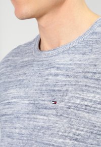 Pull texturé gris avec un col rond, présentant des variations subtiles de couleur, et un petit logo sur la poitrine en rouge, blanc et bleu.