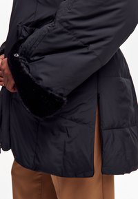 Sort pufjakke med en lynlåset sideåbning, med faux fur kant ved ærmerne og en quiltet tekstur. Blødt og isoleret materiale.