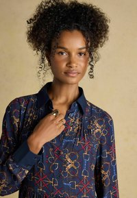 Tumesinise mustriga särk, millel on kuldsete ja punaste kujunditega, koos krae ja nööpidega esiosaga. Mudel kannab sõrmust, näidates detailseid tekstuure.