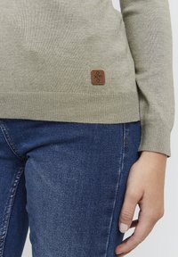 Lys grønstrikket genser med ribbestrikket kant og mansjetter, med et brunt lærlogo-merke nederst til venstre. Kombinert med mørkeblå denimjeans.