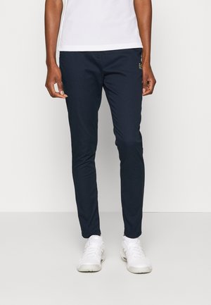 Personne portant un pantalon bleu marine slim, des baskets blanches et une chemise blanche, debout devant un fond clair uni.