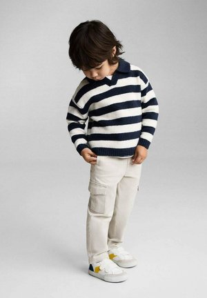 Dziecko w granatowo-białym swetrze w paski z kołnierzykiem, beżowych spodniach cargo i białych trampkach z żółtymi i niebieskimi akcentami.
