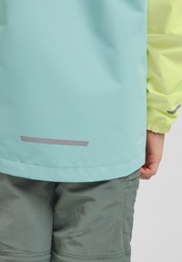 Chaqueta turquesa clara con mangas amarillas, que presenta una franja gris reflectante en el dobladillo. Tejido texturizado; combinada con pantalones cortos grises.