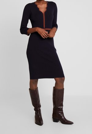 Woman in a navy knee-length dress with brown trim and three-quarter sleeves, wearing dark brown knee-high leather boots.