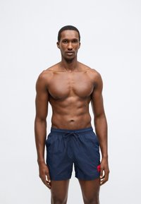 Námořnicky modré plavecké šortky z lehké látky, s elastickým pasem se stahovací šňůrkou a červenou nášivkou s logem na boku.