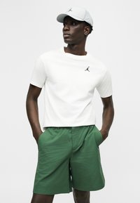 Homme noir portant un t-shirt blanc et une casquette de la marque Jordan, avec un short vert texturé, debout les mains dans les poches, regardant vers la droite.
