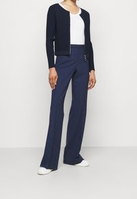 Granatowy kardigan w prążki założony na biały top z długim rękawem, zestawiony z granatowymi spodniami dzwonami i białymi sneakersami, z detalem w postaci zamka.