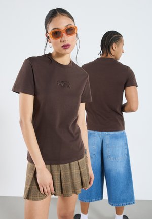 Brown cotton T-shirt featuring a small embroidered logo, paired with a pleated plaid skirt. A model wears orange sunglasses and denim shorts.
