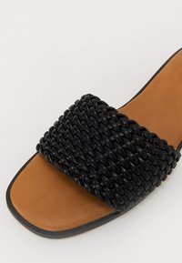 Sandale à glissière tissée noire avec un large dessus à motif texturé et une semelle lisse couleur tan, agrémentée d'un subtil bord noir.