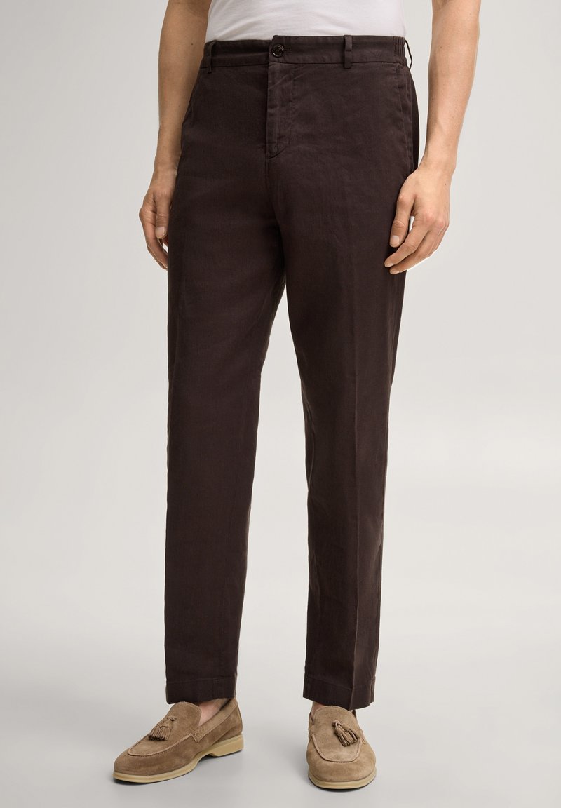 Mann trägt dunkelbraune gerade geschnittene Hose und beigefarbene Wildleder-Loafers mit Quasten, steht vor einem schlichten hellen Hintergrund.
