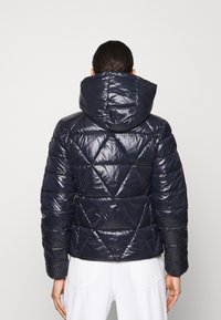 Marinblå pufferjacka med en glänsande finish, diamantquiltat mönster och huva. Har elastiska ärmslut och en figurnära silhuett.