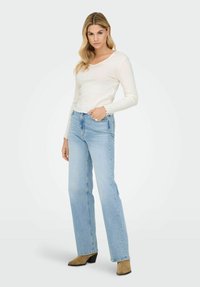 Kobieta z długimi blond włosami, ubrana w białą bluzkę z długim rękawem, jasnoniebieskie dżinsy i brązowe zamszowe botki na obcasie, stojąca z jedną ręką w kieszeni.