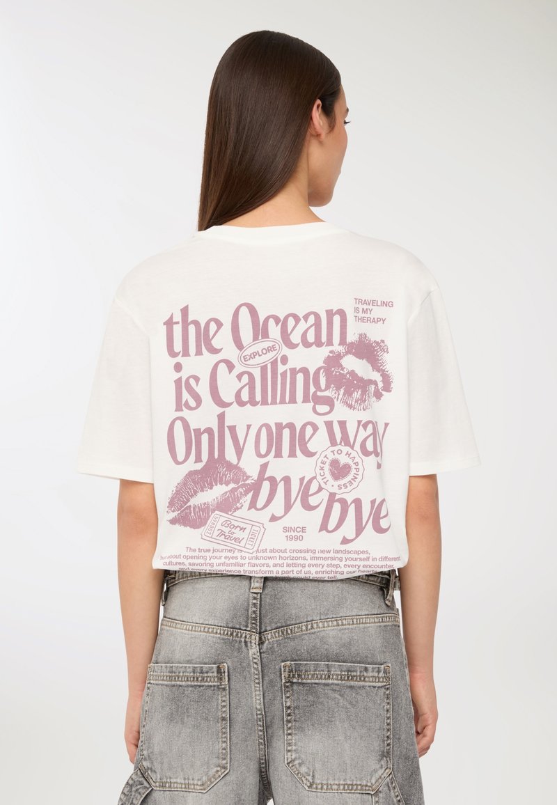 Kobieta z prostymi brązowymi włosami, nosząca białą krótką koszulkę z różowym motywem podróżniczym oraz grafiką na plecach, w połączeniu z szarymi dżinsowymi szortami.