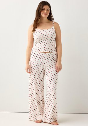 Giovane donna in piedi a piedi nudi che indossa una canotta bianca a coste e pantaloni larghi con piccole stampe floreali rosse su sfondo neutro.