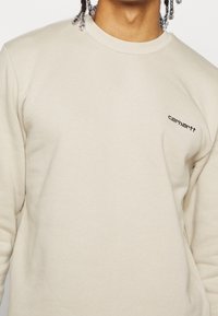 Persona que lleva un sudadera de cuello redondo Carhartt beige, mostrando la parte superior del torso y parte del cabello trenzado con cuentas.