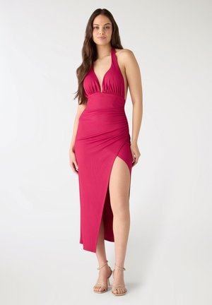 Kobieta o długich brązowych włosach, ubrana w różową, bez rękawów sukienkę na szyję z rozcięciem do uda oraz beżowe szpilki z paskiem wokół kostki.