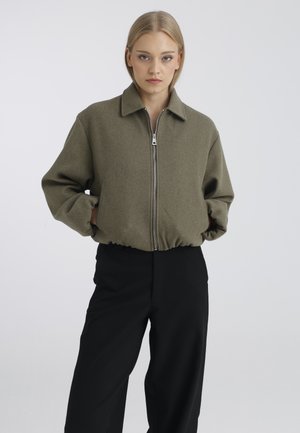 Jeune femme portant une veste zippée vert olive et un pantalon noir, debout les mains dans les poches devant un fond clair uni.