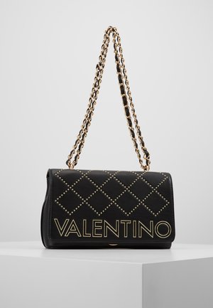 Zwarte Valentino handtas met goudkleurig spijker-diamantpatroon en gouden kettingriem, tentoongesteld op een wit voetstuk tegen een grijze achtergrond.