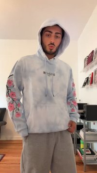 Homem com um hoodie azul claro com desenhos florais rosa nas mangas e calças de moletom cinzas, de pé dentro de casa perto de prateleiras e uma cadeira.