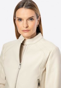 Giacca in pelle crema con colletto alto, chiusura frontale con zip e dettagli di cucitura orizzontale. Presenta due tasche laterali con zip.