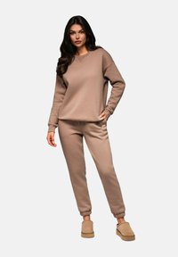 Ljusbrun sweatshirt och matchande joggers i mjukt tyg, med elastiska ärmslut och en sidetikett, tillsammans med beige sk slip-on skor.