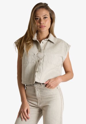 Donna con capelli lunghi castano chiaro che indossa un crop top beige senza maniche con bottoni e pantaloni coordinati a vita alta, mano in tasca.