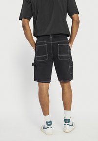 Black cargo shorts with white stitching, featuring multiple pockets on the back and sides. Worn with white Nike sneakers and ankle socks.