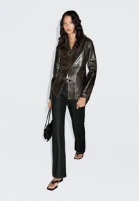 Vrouw met lang krullend haar draagt een donkerbruine leren blazer, zwarte rechte jeans, zwarte sandalen en heeft een kleine zwarte handtas bij zich.