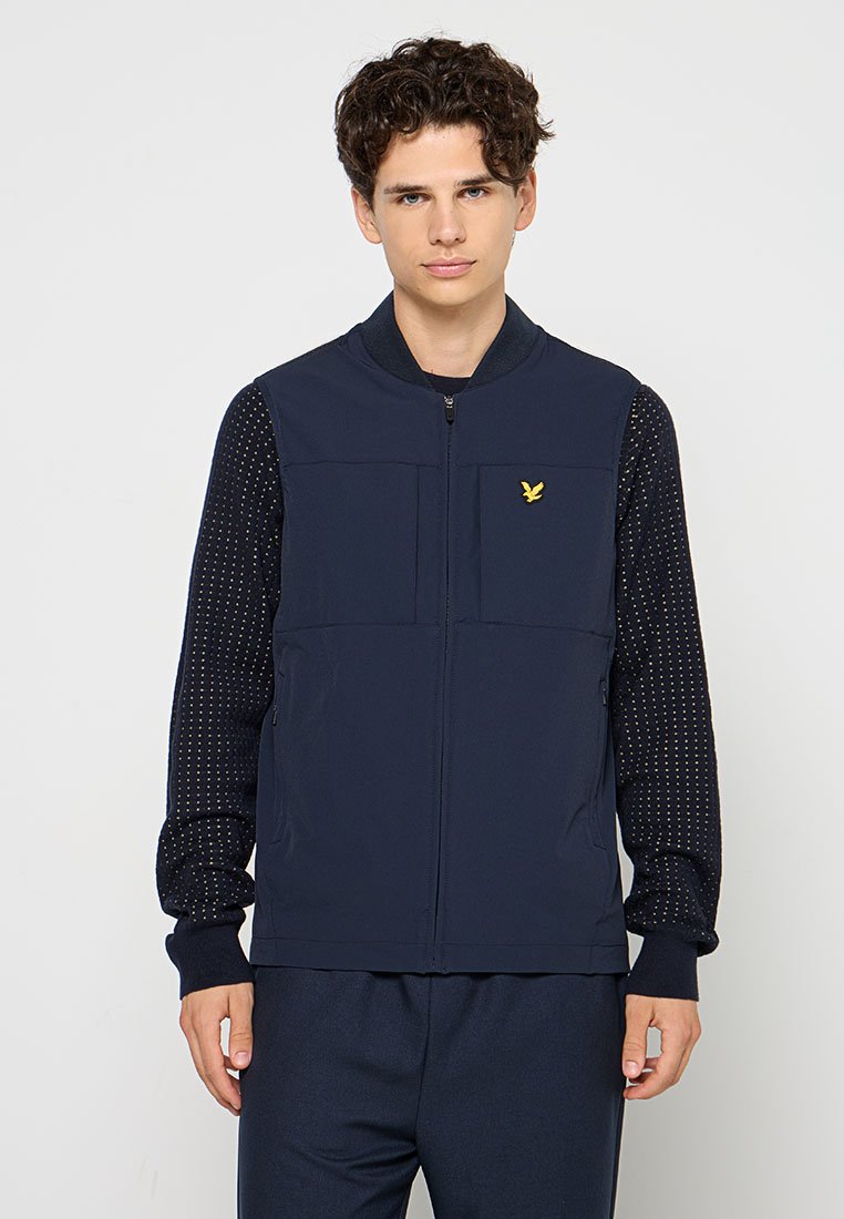 Gilet bleu marine avec deux poches avant, fermeture éclair et manches à motifs sombres. Présente un petit logo jaune sur la poitrine.