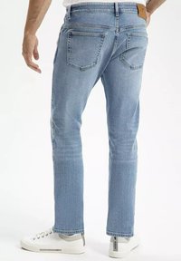 Jasnoniebieskie dżinsy z prostymi nogawkami, z dwiema tylnymi kieszeniami i beżową skórzaną łatą na pasku. Noszone z białymi sneakersami.