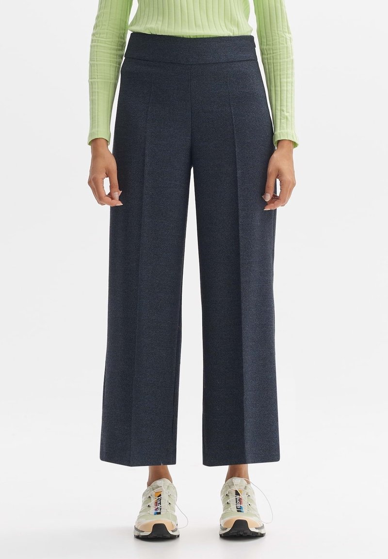 Pantalons navy à jambes larges en tissu texturé avec une taille haute, associés à des baskets légères et sportives. Les plis verticaux renforcent la structure.