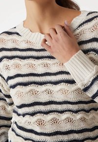 Svetr s krajkovým vzorem v krémové barvě s námořnickými pruhy. Má žebrovaný výstřih a manžety, které zdůrazňují otevřenou pletenou texturu a vlnitý design.