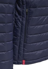 Marineblauwe gewatteerde jassectie met verticale ritszak en rode Hummel-logo label nabij de zoom.
