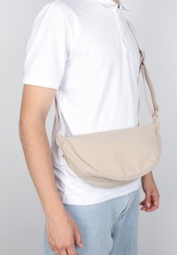 Uomo che indossa un polo bianco e jeans chiari con una borsa a tracolla beige posata in vita su uno sfondo grigio.
